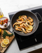 A close-up action shot of a person's hands holding a black frying pan over a countertop, tossing a colorful stir-fry of vegetables and meat. A ceramic bowl sits below, with a modern gray tiled wall in the background.