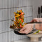 A close-up action shot of a person's hands holding a black frying pan over a countertop, tossing a colorful stir-fry of vegetables and meat. A ceramic bowl sits below, with a modern gray tiled wall in the background.