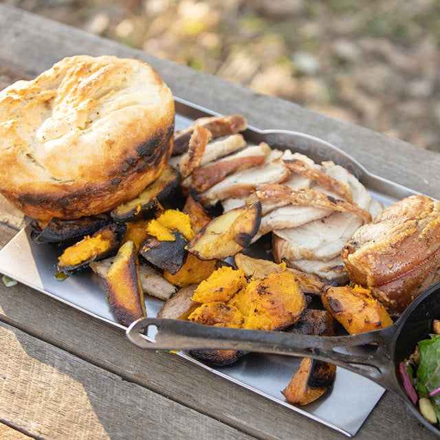 A rustic outdoor meal served on a wooden table, featuring a large golden-brown biscuit, sliced roasted meat, roasted sweet potatoes, and a side of mixed greens in a black bowl.