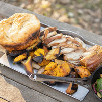 A rustic outdoor meal served on a wooden table, featuring a large golden-brown biscuit, sliced roasted meat, roasted sweet potatoes, and a side of mixed greens in a black bowl.