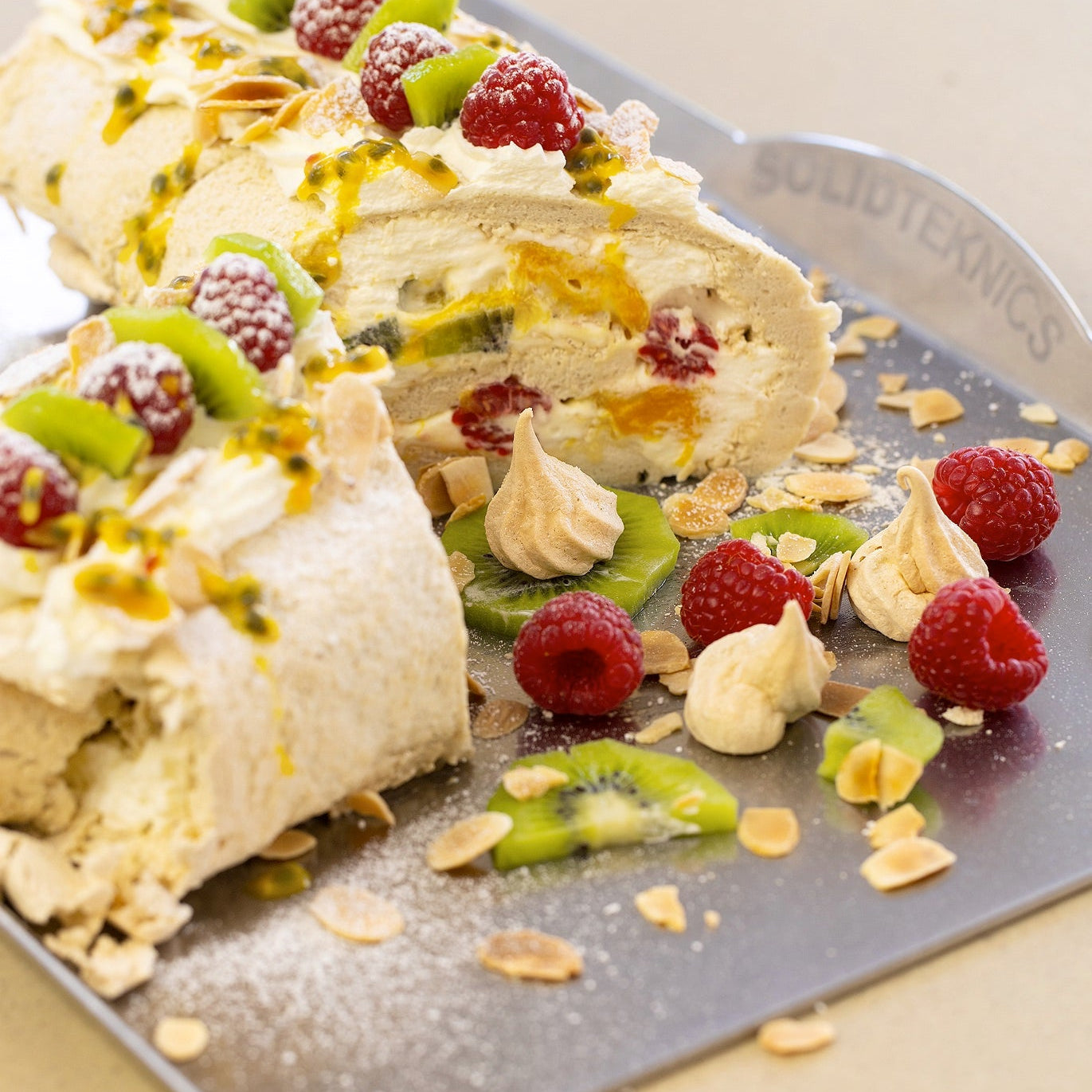 A sliced meringue cake roll decorated with fresh raspberries, kiwi slices, orange zest, and piped meringue kisses, served on a dark metal tray with a knife.