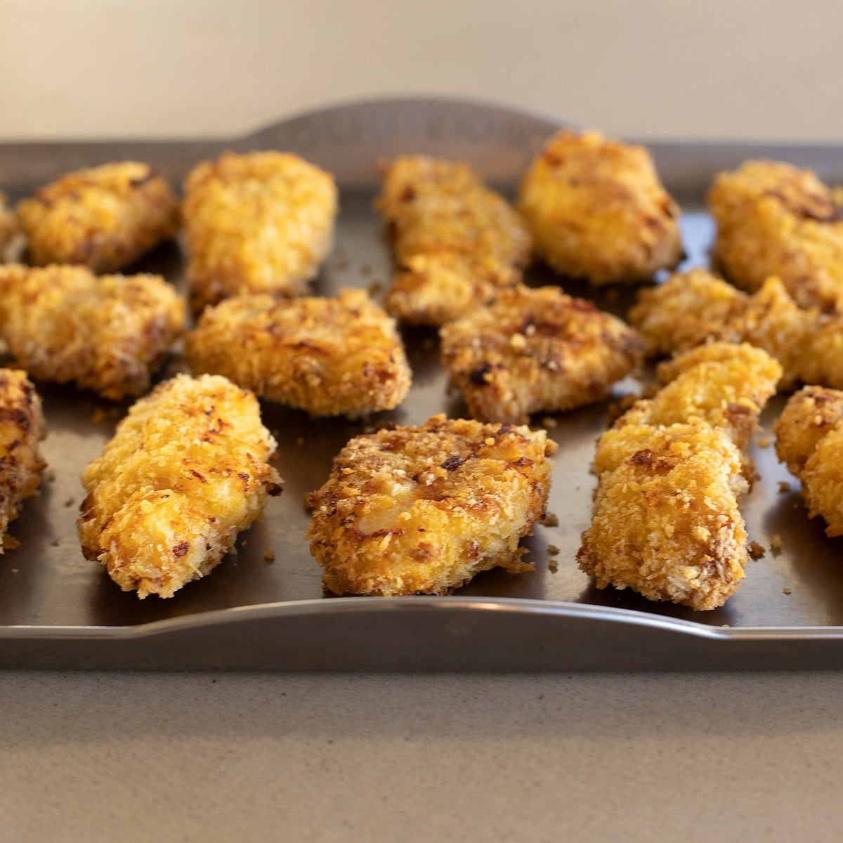 Golden-brown breaded chicken nuggets arranged on a dark metal baking sheet, photographed from above on a neutral countertop.