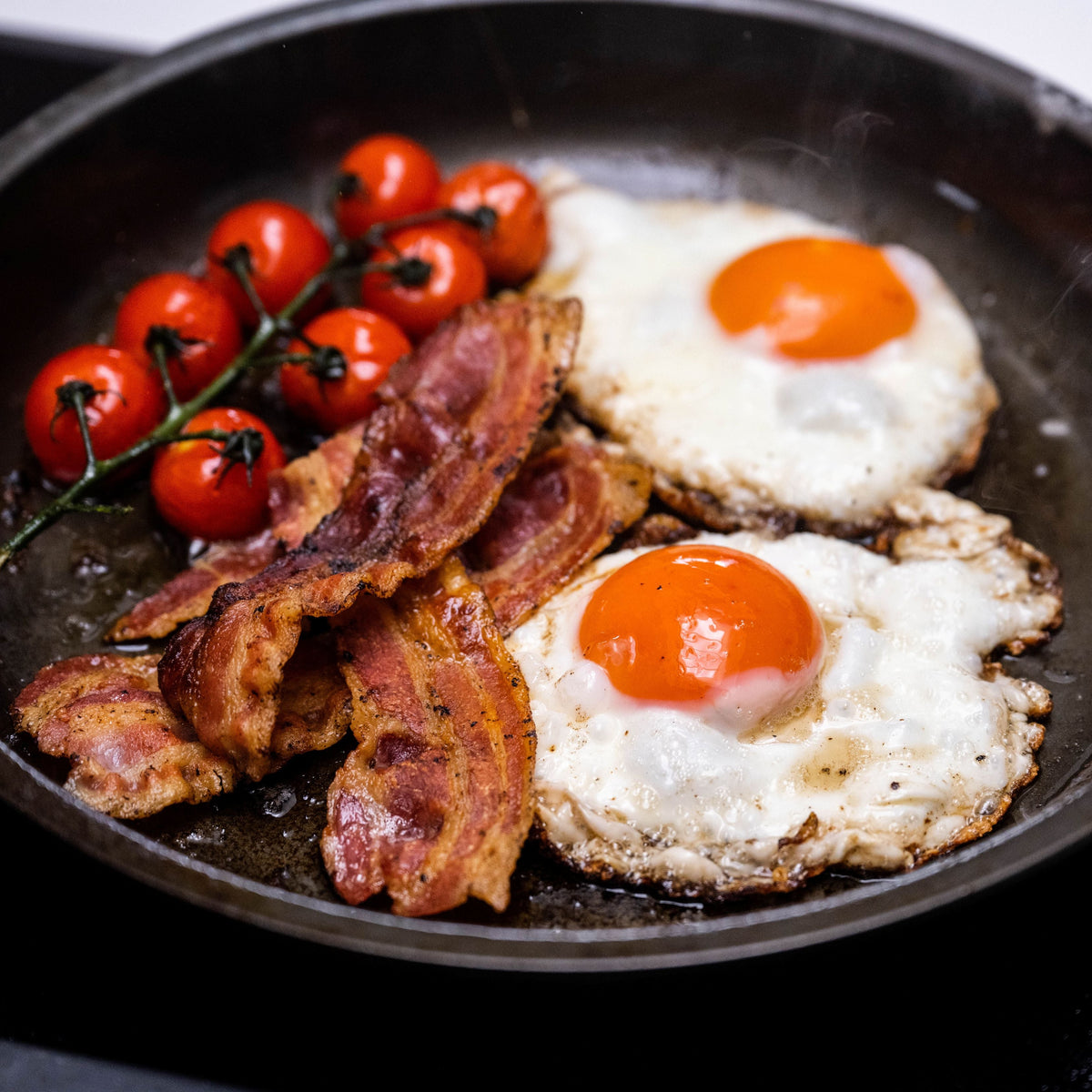 A wrought iron skillet on a black stovetop, filled with two sunny-side-up eggs, crispy bacon strips, and a cluster of red cherry tomatoes on the vine.