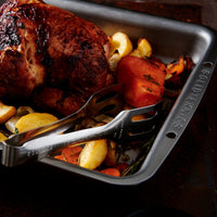 A roasted whole chicken in a dark metal baking pan with vegetables and a metal serving fork, photographed on a dark rustic surface.