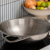 A stainless steel wok with integrated handles sits on a black induction cooktop in a modern kitchen setting, with a fruit bowl blurred in the background.