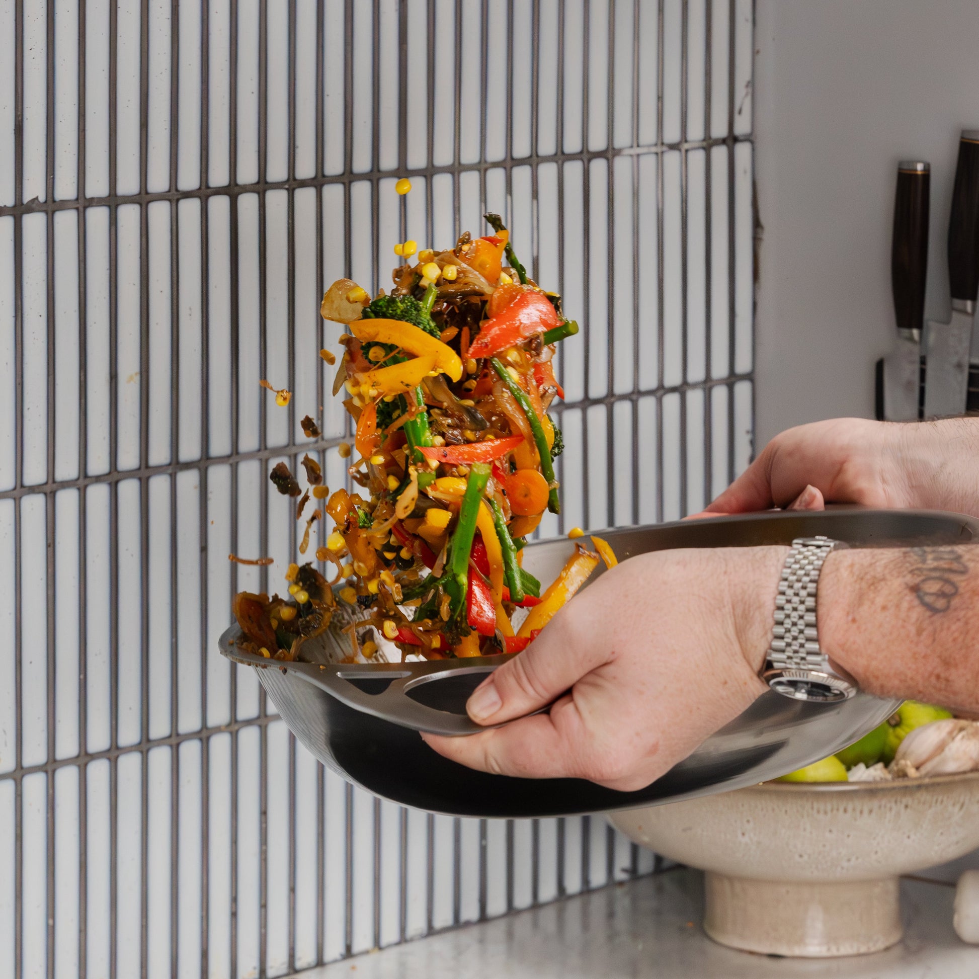 A close-up action shot of a person's hands holding a black frying pan over a countertop, tossing a colorful stir-fry of vegetables and meat. A ceramic bowl sits below, with a modern gray tiled wall in the background.